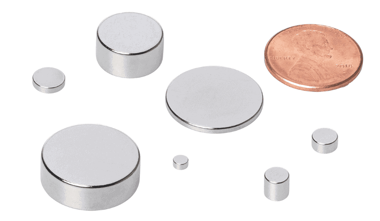 Assorted small silver neodymium disc magnets of different sizes arranged on a white surface, with a U.S. penny beside them.
